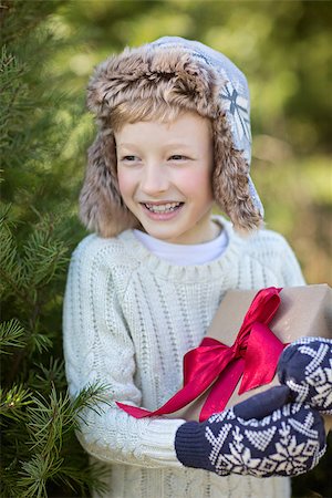 snow cosy - beautiful smiling boy in trapper hat and mittens holding christmas gift standing by the tree, winter holiday concept Stock Photo - Budget Royalty-Free & Subscription, Code: 400-09082513