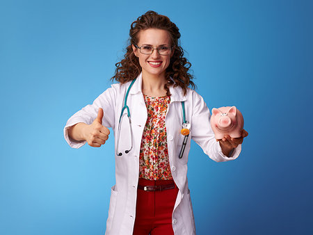 happy paediatrist woman in white medical robe with piggy bank showing thumbs up against blue background Stock Photo - Budget Royalty-Free & Subscription, Code: 400-09237713