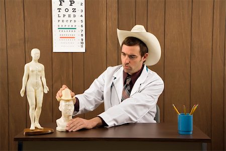 simsearch:400-03924532,k - Mid-adult Caucasian male doctor wearing cowboy hat sitting at desk playing with figurine. Stock Photo - Budget Royalty-Free & Subscription, Code: 400-03924534