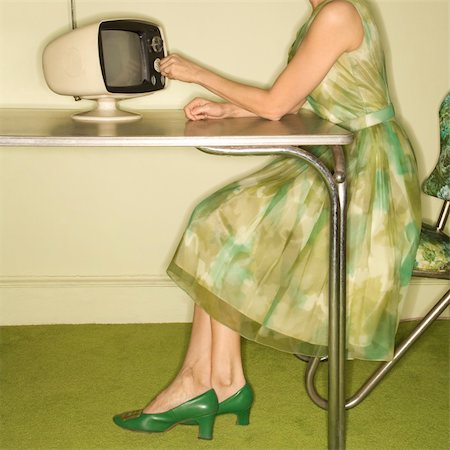 Side view of Caucasian mid-adult woman wearing green vintage dress sitting at 50's retro dinette set turning old televsion knob. Stockbilder - Microstock & Abonnement, Bildnummer: 400-03925729