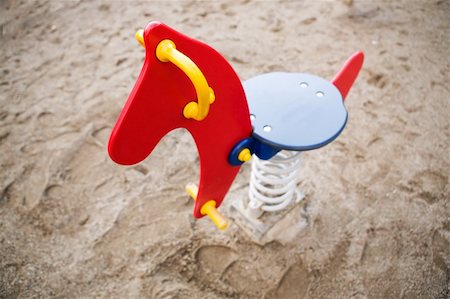 A brightly colored red, blue and yellow rocking horse in a public playground surrounded by sand. - focus on the head with a shallow depth of field. Foto de stock - Super Valor sin royalties y Suscripción, Código: 400-03938786