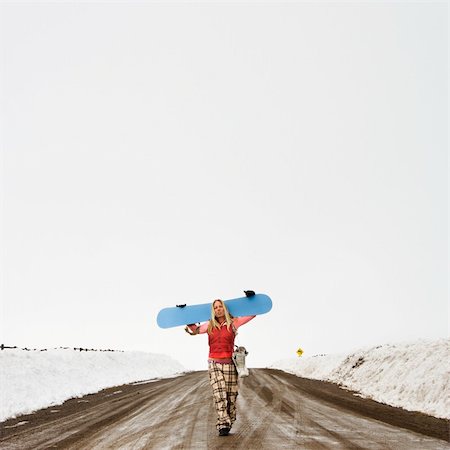 simsearch:400-03944669,k - Young woman in winter clothes walking alone down muddy dirt road carrying snowboard and boots. Photographie de stock - Aubaine LD & Abonnement, Code: 400-03943524