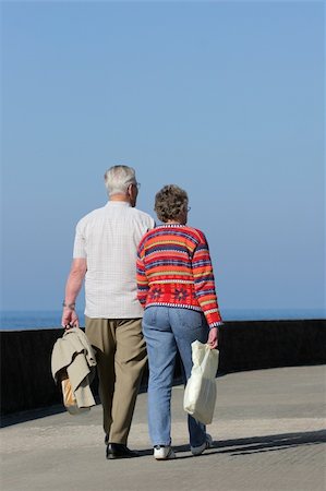 simsearch:400-05711437,k - Two elderly people walking together on a beach promenade, holding shopping bags, with the female wearing a vivid colored jumper. Blue sky and sea (out of focus) to the rear. Stock Photo - Budget Royalty-Free & Subscription, Code: 400-03953219