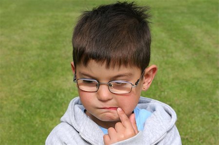 photographic portraits poor people - Young boy wearing glasses with a thoughtful expression on his face and a forefinger to his mouth. Grass (out of focus) to the rear. Stock Photo - Budget Royalty-Free & Subscription, Code: 400-03987177