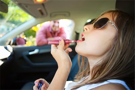 rossetto - Young girl applying lip gloss pretending to be old enough to drive as her smiling father watches on on a sunny summer evening in Portland, Oregon, USA Fotografie stock - Premium Royalty-Free, Codice: 600-06531444