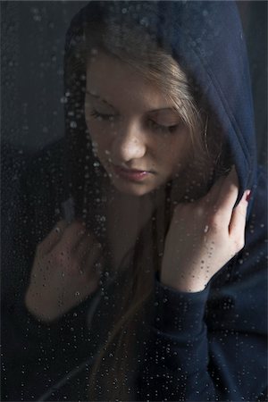 simsearch:600-06553417,k - Portrait of young woman behind window, wet with raindrops, wearing hoodie, looking down Stock Photo - Premium Royalty-Free, Code: 600-06786760