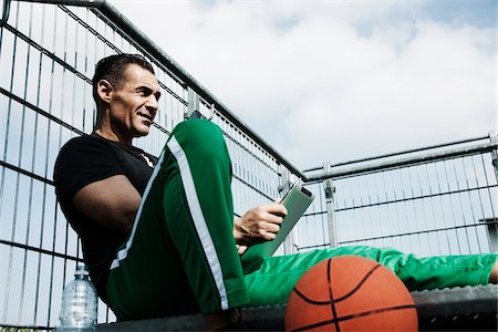fence (enclosure) - Mature man sitting at top of stairs on outdoor basketball court looking at tablet computer, Germany Stock Photo - Premium Royalty-Free, Code: 600-06786852