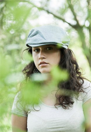 Close-up portrait of teenaged girl wearing cap outdoors, looking at camera through leaves, Germany Stock Photo - Premium Royalty-Free, Code: 600-06899851