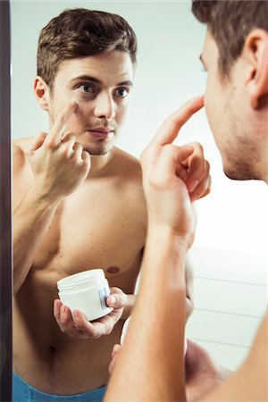 Close-up of young man reflected in bathroom mirror, holding jar of cream and applying to face, studio shot Stock Photo - Premium Royalty-Free, Code: 600-07278944