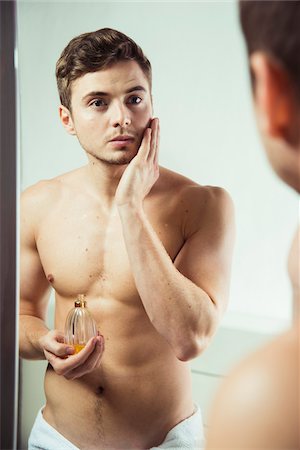 putting on - Young man looking in bathroom mirror, applying cologne to face, studio shot Stock Photo - Premium Royalty-Free, Code: 600-07278947