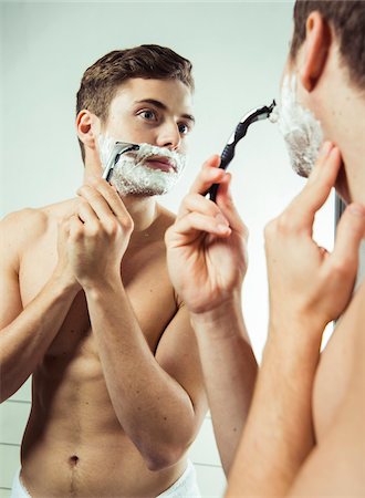 Young man looking in bathroom mirror, shaving with razor, studio shot on white background Foto de stock - Sin royalties Premium, Código: 600-07278955