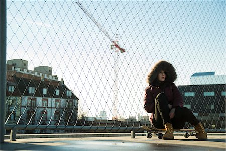 Teenage girl wearing winter coat, sitting on skateboard outdoors, next to chain link fence near comercial dock, Germany Foto de stock - Sin royalties Premium, Código: 600-07310998