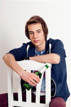 Portrait of teenage boy sitting on chair holding bottle of beer, smiling and looking at camera, studio shot on white background Foto de stock - Sin royalties Premium, Código: 600-07311017