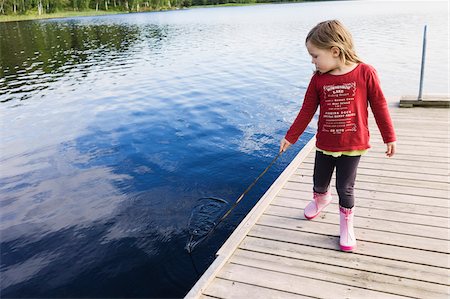 3 year old girl in red shirt on a pier holding a stick and playing in the water, Sweden Stock Photo - Premium Royalty-Free, Code: 600-07311126