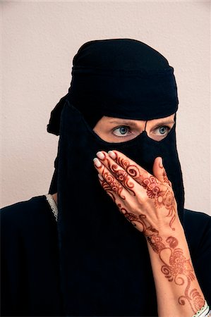 Close-up portrait of woman wearing black muslim hijab and muslim dress, looking to the side with hand covering mouth and showing arms and hands painted with henna in arabic style, studio shot on whtie background Stock Photo - Premium Royalty-Free, Code: 600-07434939