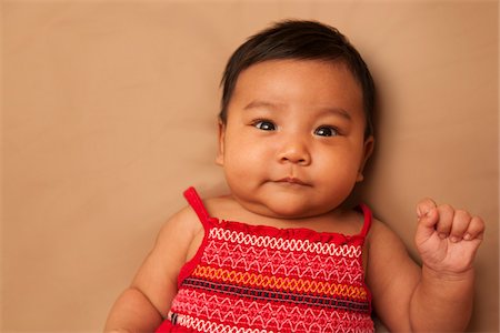 simsearch:600-07453955,k - Close-up portrait of Asian baby lying on back, wearing red dress, looking at camera and smiling, studio shot on brown background Stock Photo - Premium Royalty-Free, Code: 600-07453956