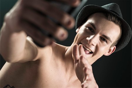 simsearch:600-06505868,k - Close-up portrait of young man wearing fedora, taking a selfie with cellphone, studio shot on black background Foto de stock - Sin royalties Premium, Código: 600-07567382