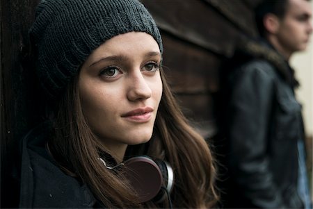 Close-up portrait of teenage girl outdoors, wearing hat and headphones around neck, with young man in background, Germany Stock Photo - Premium Royalty-Free, Code: 600-07567383