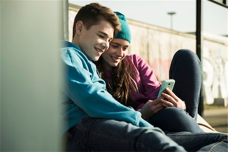 Teenage girl and boy sitting on ground, smiling and looking at cell phone together, Mannheim, Germany Stock Photo - Premium Royalty-Free, Code: 600-07584764