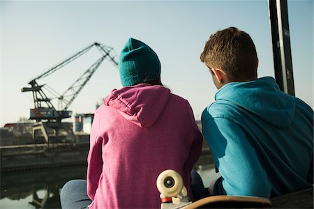 Backview of teenage girl and boy sitting on bench outdoors with skateboard, industrial area, Mannheim, Germany Foto de stock - Sin royalties Premium, Código: 600-07584763