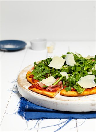 Pizza with Ham, Arugula and Parmisan Cheese on Wooden Cutting Board on Napkin Stock Photo - Premium Royalty-Free, Code: 600-08060051