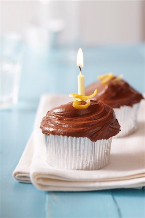 Chocolate frosted cupcakes with lemon zest and a lit birtday candle on a blue background Stock Photo - Premium Royalty-Free, Code: 600-09119367