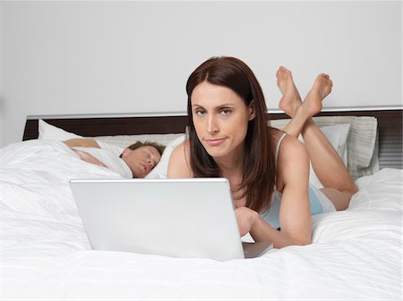 simsearch:600-01787405,k - Portrait of Woman Using Laptop Computer in Bed while Man Sleeps Stock Photo - Premium Royalty-Free, Code: 600-01295821