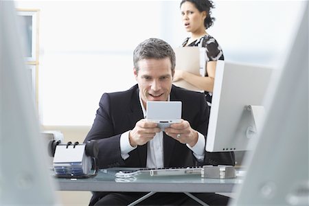 simsearch:600-02081724,k - Co-worker looking at Businessman Playing Handheld Video Game at Desk Stock Photo - Premium Royalty-Free, Code: 600-02081702