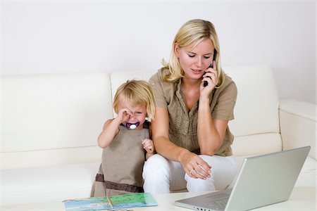 Mother Using Laptop and Cell Phone, Toddler Standing Beside Her Crying Foto de stock - Sin royalties Premium, Código: 600-02332632