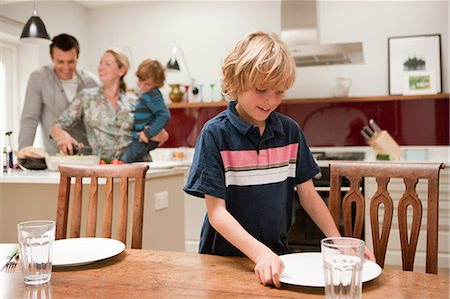 Son helping to lay table with parents and brother visible behind in kitchen Stock Photo - Premium Royalty-Free, Code: 614-05650637