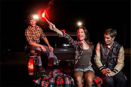 Three friends sitting on tailgate of car at night, girl holding sparkler Stock Photo - Premium Royalty-Free, Code: 614-06402549