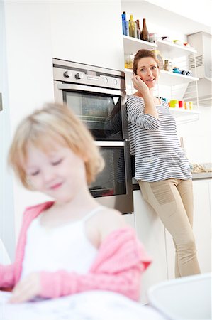 people candid happy - Mother on cell phone in kitchen with daughter in foreground Stock Photo - Premium Royalty-Free, Code: 614-06442567