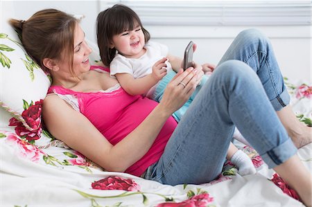 Portrait of pregnant woman and toddler daughter lounging on bed playing with smartphone Stock Photo - Premium Royalty-Free, Code: 614-06813761