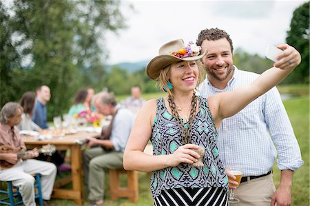 Man and woman taking self portrait with smartphone while family friends sit at table behind, outdoors Stock Photo - Premium Royalty-Free, Code: 614-07806384