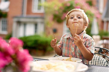 simsearch:614-03020295,k - Boy sitting at garden table sucking up spaghetti looking at camera Fotografie stock - Premium Royalty-Free, Codice: 614-08270453