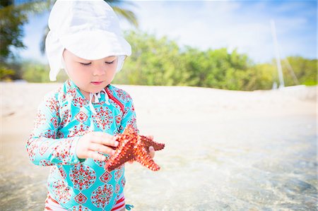 Girl on beach wearing swimear and sunhat holding starfish looking down, St. Croix, US Virgin Islands Stock Photo - Premium Royalty-Free, Code: 614-08307900