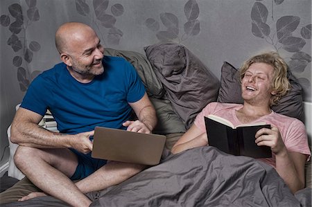person being lazy on computer - Male couple sitting up in bed laughing whilst reading and using laptop Stock Photo - Premium Royalty-Free, Code: 614-08329489