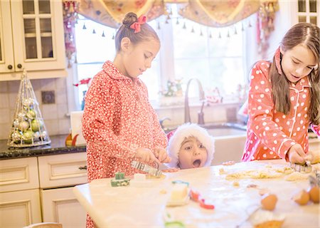 Boy in kitchen wearing santa hat making cookies with sisters peeking over kitchen counter open mouthed Foto de stock - Sin royalties Premium, Código: 614-08392735