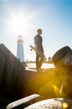 simsearch:614-08081220,k - Young man holding skateboard, standing on breakwater near lighthouse Fotografie stock - Premium Royalty-Free, Codice: 614-08487827