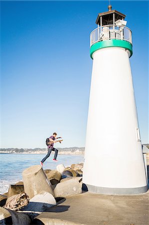 simsearch:614-08487824,k - Young man holding skateboard, jumping off of breakwater, beside lighthouse Foto de stock - Sin royalties Premium, Código: 614-08487830