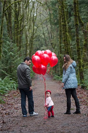 Portrait of female toddler holding a bunch of red balloons with parents in forest Stock Photo - Premium Royalty-Free, Code: 614-08578840