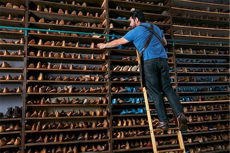 Male cobbler in traditional shoe shop on ladder selecting shoe last Foto de stock - Royalty Free Premium, Número: 614-08641735