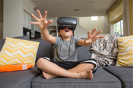 play with kid - Young boy sitting cross legged on sofa, wearing virtual reality headset, hands reaching out in front of him Stock Photo - Premium Royalty-Free, Code: 614-08884615