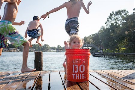 simsearch:640-02657491,k - Young girl hiding behind bucket on jetty, children jumping into lake Stock Photo - Premium Royalty-Free, Code: 614-09038554