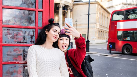 Two young stylish women taking smartphone selfie by red phone box, London, UK Foto de stock - Sin royalties Premium, Código de la imagen: 614-09057108