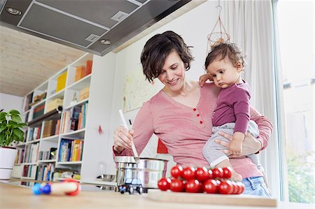 Mother holding baby girl in kitchen, whilst stirring pot on stove Stock Photo - Premium Royalty-Free, Code: 614-09127305