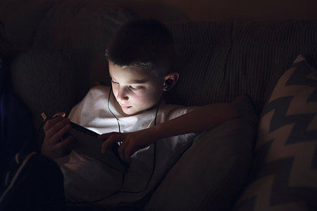 Boy on sofa wearing earphones looking down using digital tablet Stock Photo - Premium Royalty-Free, Code: 614-09212426