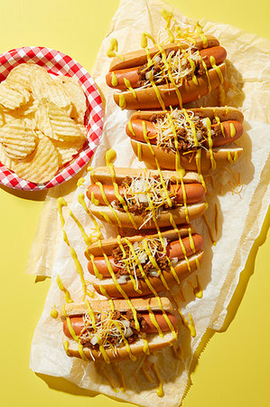 simsearch:6102-07843397,k - Row of coney hotdogs with drizzled mustard and plate of potato chips on yellow background, overhead view Photographie de stock - Premium Libres de Droits, Code: 614-09276792