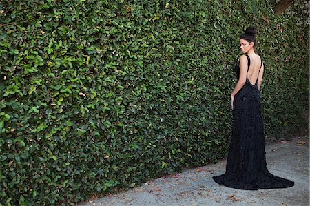 Young woman wearing black evening dress, standing by hedge Stock Photo - Premium Royalty-Free, Code: 614-03763893