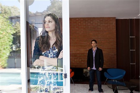 Woman looking through window with arms crossed, man in background Stock Photo - Premium Royalty-Free, Code: 614-03818619